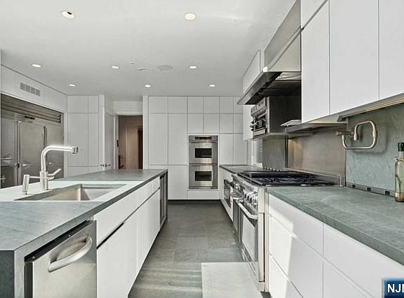 The chef's kitchen with white minimalist cabinetry, gray stone countertops, a professional six-burner gas range with double oven, a deep island sink, and stainless built-in refrigeration