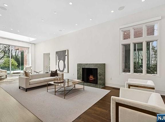 A detail of the marble fireplace at the heart of the great room, the fire lit, framed by dark hardwood floors and a glass wall opening to the backyard beyond